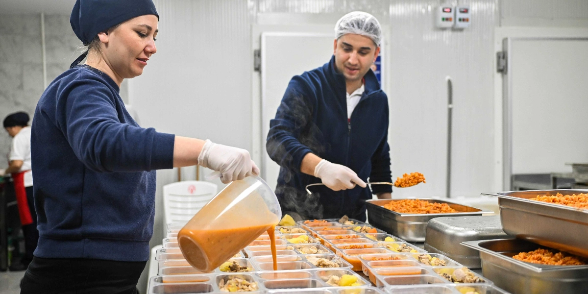 Endüstriyel bir mutfakta, başlarında bone takan bir kadın ve bir erkek çalışan, plastik yemek kaplarına servis yapıyor. Kadın çalışan elindeki sürahiden kaplara çorba doldururken, yanındaki erkek çalışan kepçeyle makarna paylaştırıyor. Önlerinde çok sayıda bölmeli yemek kabı ve büyük metal kaplarda hazırlanan yemekler bulunuyor.