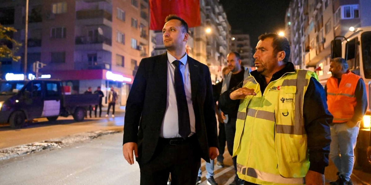 Gece vakti Bornova sokaklarında, üzerinde Bornova Belediyesi logolu fosforlu sarı bir yelek bulunan bir çalışan, takım elbiseli bir yetkiliye eliyle işaret ederek bilgi veriyor. Arka planda belediye araçları, apartmanlar ve sokak aydınlatmaları görülüyor.
