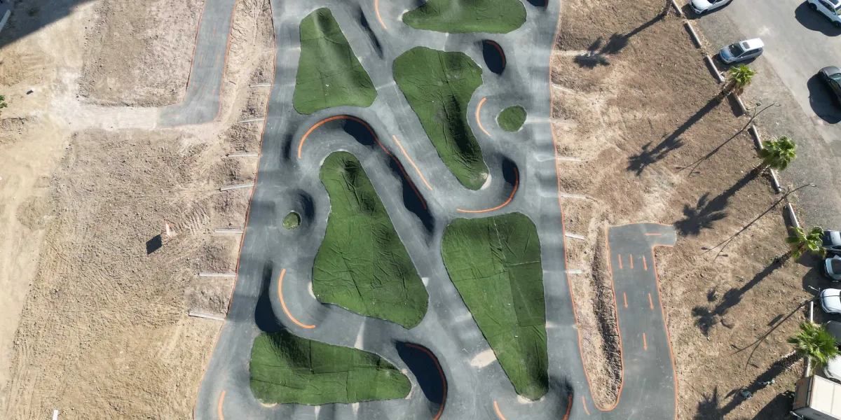 Bornova'da bulunan, bisiklet ve kaykay kullanımı için tasarlanmış, kıvrımlı asfalt yollara ve yeşil suni çim kaplı tümseklere sahip kuş bakışı bir pumptrack parkuru. Parkurun çevresi toprak zeminle çevrili olup sağ tarafta ağaçlar ve park halindeki araçlar görülmektedir.