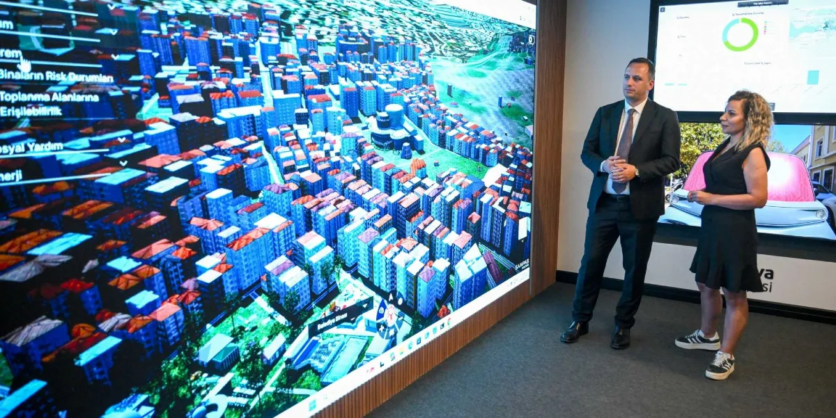 Modern bir ofis ortamında, devasa bir dokunmatik ekranın önünde duran bir erkek ve bir kadın, Bornova'nın üç boyutlu dijital ikiz modelini inceliyor. Ekranda binaların risk durumları, toplanma alanları ve sosyal yardım gibi verilerin yer aldığı bir arayüz ile şehrin kuş bakışı görünümü yer alıyor. Takım elbiseli erkek ve siyah elbiseli kadın, ekran üzerindeki verileri değerlendirirken yan taraftaki daha küçük bir ekranda ise grafikler ve bir araç görseli bulunuyor.