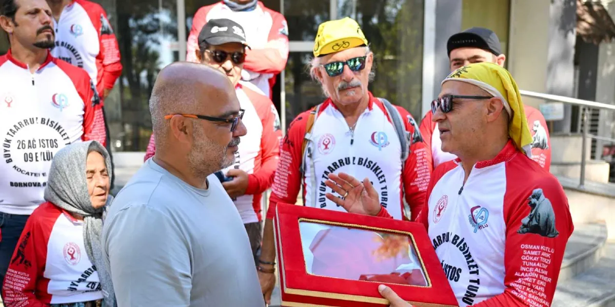Büyük Taarruz Bisiklet Turu formaları giymiş bir grup katılımcı, ellerinde kırmızı kadife kaplı, cam bölmeli bir çerçeve ile duruyor. Ön planda gri tişörtlü bir adam, elinde çerçeveyi tutan bisikletçiyle sohbet ediyor. Grubun arasında gri başörtülü yaşlı bir kadın, sarı şapkalı ve güneş gözlüklü bisikletçiler yer alıyor. Formaların üzerinde 