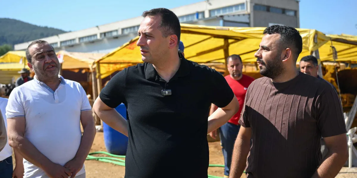 Açık havada, sarı brandalı çadırların bulunduğu bir alanda duran üç erkekten ortadaki siyah polo yaka tişörtlü kişi, yanındaki kahverengi tişörtlü kişiyle konuşuyor. Sol tarafta ise beyaz polo yaka tişörtlü bir başka kişi onları dinliyor. Arka planda ise kırmızı tişörtlü bir başka kişi daha görülüyor.