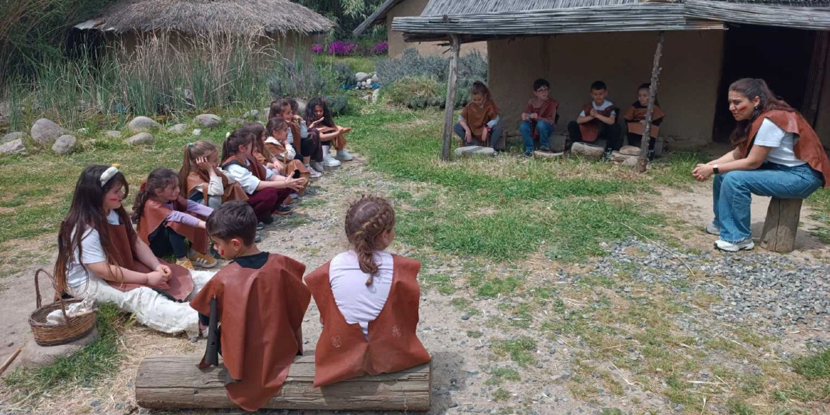 Taş Devri konseptli bir açık hava müzesinde, üzerlerinde dönem kıyafetlerini andıran kahverengi deri yelekler bulunan bir grup çocuk, eğitmenlerini dinlemek üzere çimlerin üzerinde ve kütüklerin üzerinde oturuyor. Eğitmen, çocukların karşısında bir kütüğün üzerinde oturarak onlara bir şeyler anlatıyor; arka planda ise sazdan çatılı, kerpiç görünümlü tarih öncesi dönem evleri yer alıyor.
