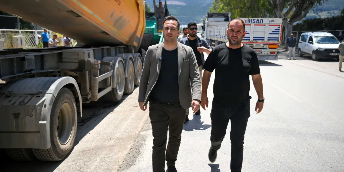 Pınar Caddesi'ndeki yol yenileme çalışmaları sırasında, gri ceketli ve siyah tişörtlü iki adam yan yana yürürken görülüyor. Arka planda damperli bir kamyon, iş makineleri ve çalışma alanında bulunan diğer görevliler yer alıyor.