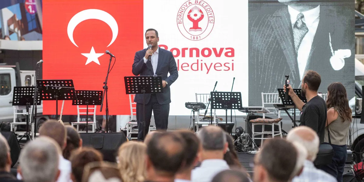 Bornova Belediye Başkanı, arkasında Türk bayrağı ve Bornova Belediyesi logosunun bulunduğu bir sahnede mikrofonla konuşma yapıyor. Sahnenin sağ tarafında Mustafa Kemal Atatürk'ün silüetini andıran büyük bir görsel yer alıyor. Ön planda ise konuşmayı dinleyen kalabalık bir izleyici grubu ve çekim yapan bir kişi bulunuyor.