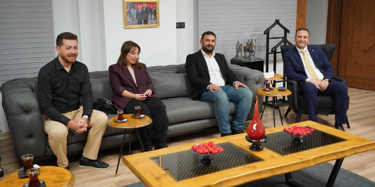 Modern bir ofis ortamında gri bir koltukta yan yana oturan üç kişi ve tekli koltukta oturan bir kişi, gülümseyerek kameraya bakıyor. Soldan sağa doğru; siyah gömlekli ve bej pantolonlu bir erkek, bordo ceketli bir kadın, beyaz gömlek ve siyah ceketli, kot pantolonlu bir erkek ile sağ tarafta lacivert takım elbiseli ve sarı kravatlı bir erkek yer alıyor. Önlerinde ahşap bir orta sehpa üzerinde kırmızı süslemeler ve dekoratif bir vazo bulunuyor. Yan sehpalarda ise ince belli bardaklarda çaylar servis edilmiş durumda.