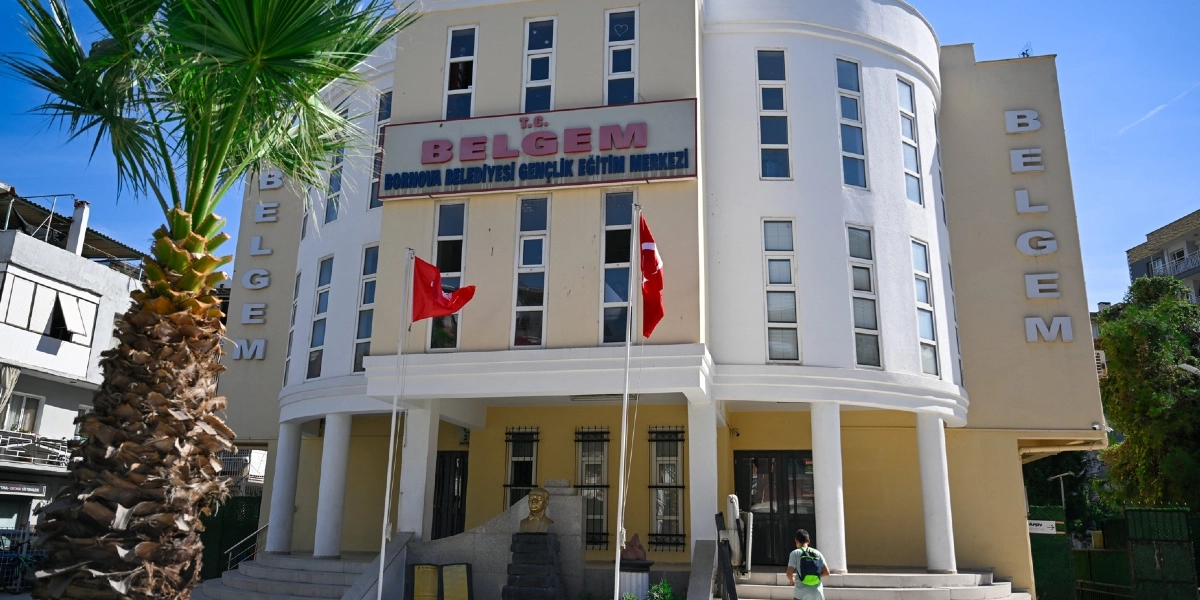Açık renkli, modern mimariye sahip Bornova Belediyesi Gençlik Eğitim Merkezi (BELGEM) binasının dış görünüşü. Binanın ön cephesinde büyük harflerle 