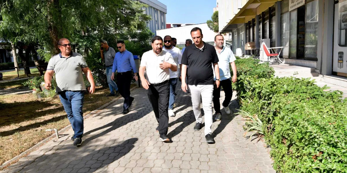Belediye başkanı ve beraberindeki heyet, Fincancılar Çarşısı'nın dışındaki taş döşeli yolda yan yana yürüyerek ilerliyor. Başkan beyaz gömlek ve siyah pantolon giymiş, yanındaki siyah tişörtlü kişiyle sohbet ediyor. Grubun geri kalanı farklı renklerde tişört ve gömleklerle onlara eşlik ediyor. Sağ tarafta dükkanların önünde yeşil bitkiler, sol tarafta ise ağaçlıklı bir park alanı bulunuyor.