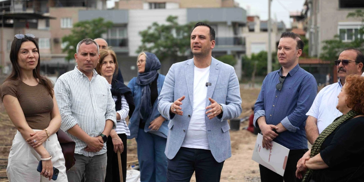 Açık havada, toprak bir alanda bir grup insan bir yetkilinin konuşmasını dinliyor. Ortada duran ve açık mavi bir ceket giyen kişi, elleriyle jest yaparak bir şeyler anlatıyor. Grubun geri kalanı dikkatle onu dinlerken, sağ tarafta duran kişi elinde üzerinde Bornova Belediyesi logosu bulunan bir dosya tutuyor. Arka planda ise modern apartmanlar ve ağaçlar görülüyor.