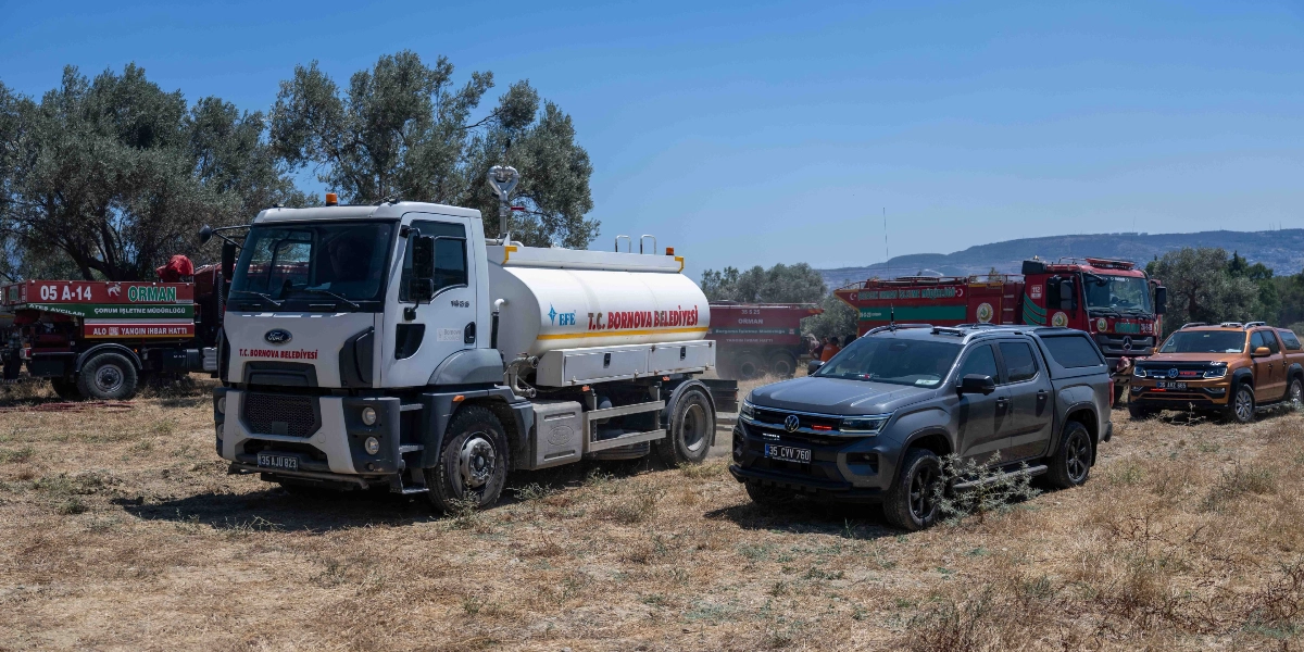 Homeros Vadisi'ndeki yangın bölgesinde, üzerinde T.C. Bornova Belediyesi yazan beyaz bir su tankeri, gri bir Volkswagen Amarok arazi aracı ve arka planda Orman Genel Müdürlüğü'ne ait kırmızı itfaiye araçları ile diğer hizmet araçları kuru otlarla kaplı bir arazide yan yana duruyor.