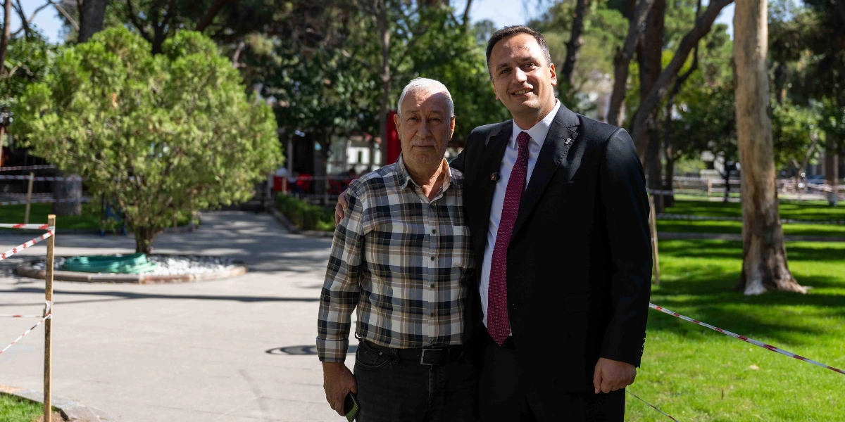 Yeşil alanlı bir parkta, yan yana duran iki erkekten biri kareli gömlek giymiş, diğeri ise koyu renkli bir takım elbise ve bordo kravat ile poz veriyor. Arka planda ağaçlar ve çevresi şeritlerle çevrilmiş bir bahçe düzenlemesi görülüyor.