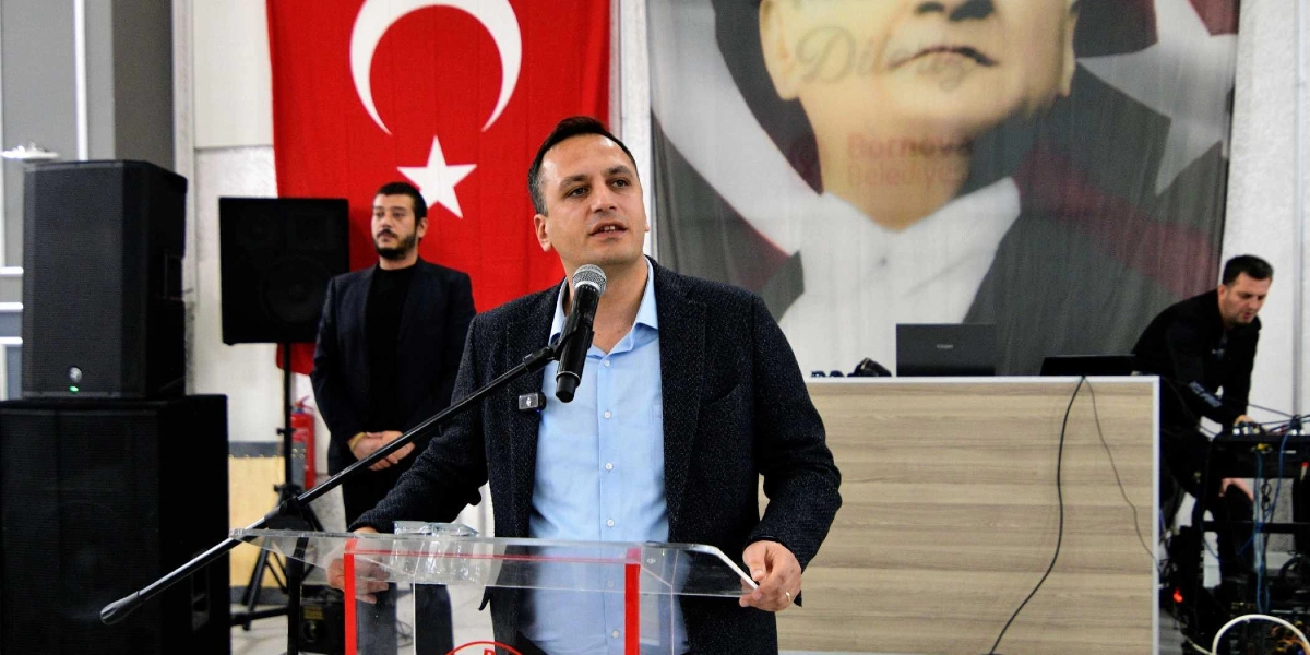 Bornova Belediye Başkanı Ömer Eşki, bir kürsü arkasında mikrofonla konuşma yapıyor. Arka planda Türk bayrağı ve Atatürk posteri yer alırken, yanında bir görevli ve sağ tarafta ses sistemini yöneten bir kişi bulunuyor.
