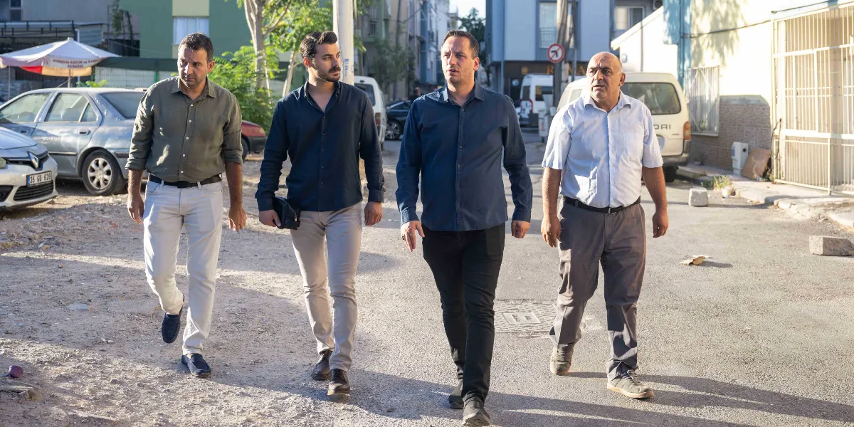 Bornova'da bir sokakta yan yana yürüyen dört erkek. Soldan sağa doğru; haki renkli gömlek ve açık renkli pantolon giyen, lacivert gömlek ve bej pantolon giyen, koyu mavi gömlek ve siyah pantolon giyen ve son olarak beyaz kısa kollu gömlek ile gri pantolon giyen dört kişi, güneşli bir havada sohbet ederek ilerliyor. Arka planda park halindeki araçlar ve binalar yer alıyor.