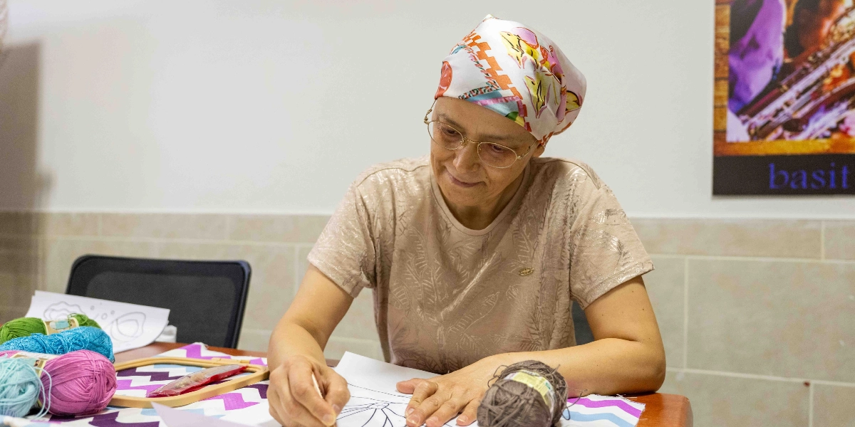 Başında renkli desenli bir eşarp ve gözlük takan orta yaşlı bir kadın, masanın başında el sanatları çalışması yapıyor. Kadın, önündeki kağıda çizim yaparken gülümsüyor; masanın üzerinde ise nakış kasnağı ve çeşitli renklerde yumak iplikler bulunuyor.