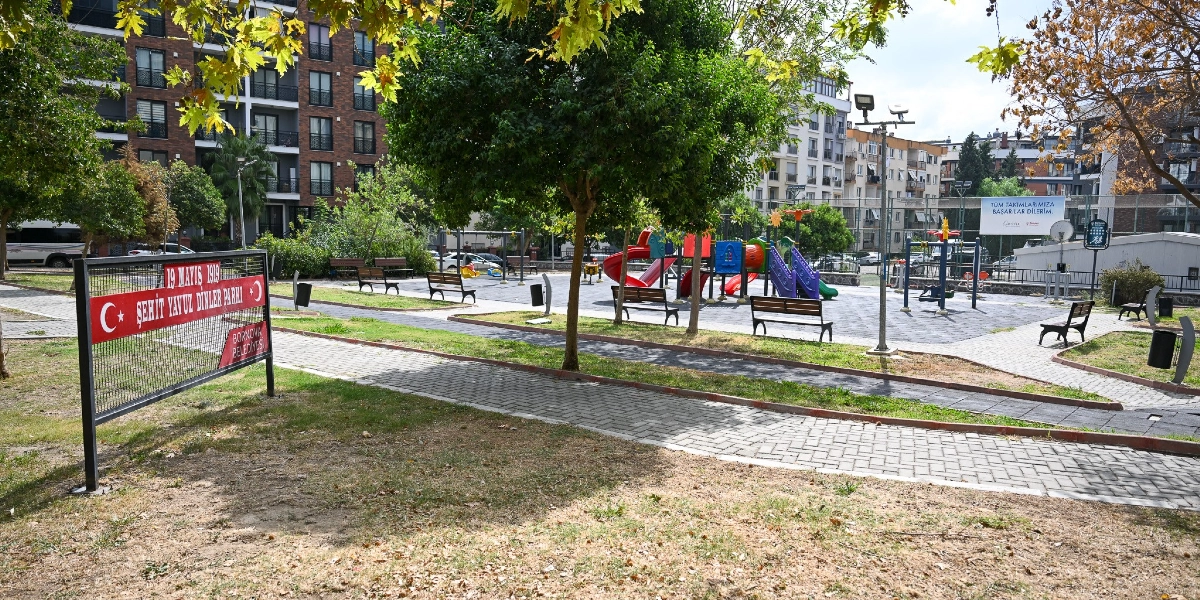 Bornova Belediyesi'ne ait 19 Mayıs 1919 Şehit Yavuz Dinler Parkı'nın genel görünümü. Parkın girişinde üzerinde parkın ismi ve Türk bayrakları bulunan bir tabela yer alıyor. Parkın içinde çocuk oyun grubu, banklar, yürüyüş yolları ve ağaçlar bulunuyor. Arka planda ise çok katlı apartmanlar ve spor sahası görülüyor.
