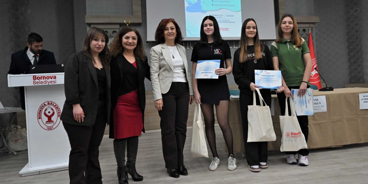 Bornova Belediyesi tarafından düzenlenen bir etkinlikte, ellerinde katılım belgeleri ve bez çantalar bulunan üç genç kız öğrenci ile onlara eşlik eden üç yetişkin kadın yan yana duruyor. Arka planda Bornova Belediyesi logosunun yer aldığı bir kürsü, üzerinde 