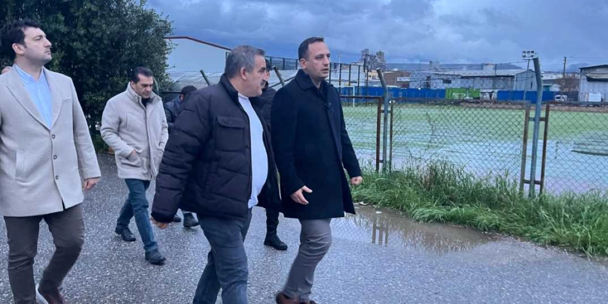 Bornova Belediye Başkanı Ömer Eşki ve beraberindeki heyet, Doğanlar Mahallesi'ndeki bir spor sahasının yanında incelemelerde bulunuyor. Dört kişilik grup, asfalt bir yolda yürürken saha hakkında konuşuyor. Arka planda tel örgülerle çevrili yeşil bir saha ve sanayi yapıları görülüyor.