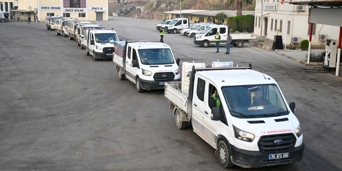 Bornova Belediyesi'ne ait beyaz renkli kamyonetler, arkalarında yeni çöp konteynerleri yüklü halde bir şantiye alanında sıra halinde bekliyor. Arka planda üzerinde 