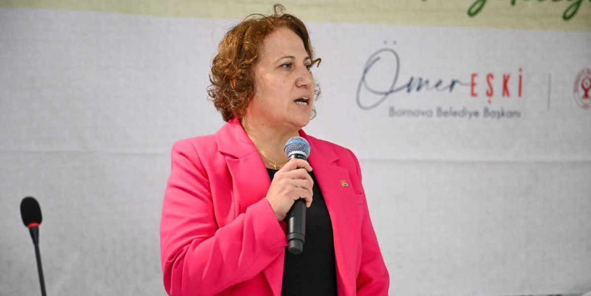 Kıvırcık kahverengi saçlı, pembe ceketli bir kadın, elinde tuttuğu mikrofonla konuşma yapıyor. Arka planda üzerinde Ömer Eşki Bornova Belediye Başkanı yazılı beyaz bir pankart yer alıyor.