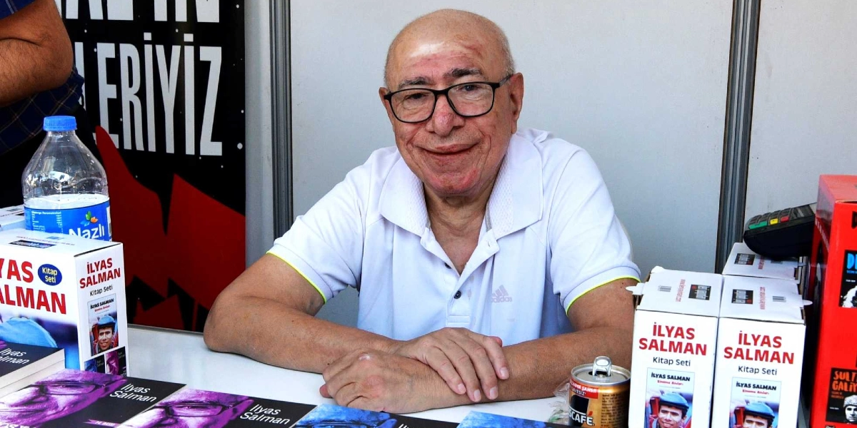 Beyaz polo yaka tişört giymiş, gözlüklü İlyas Salman, önünde kendi kitap setlerinin bulunduğu bir masada oturarak gülümseyerek kameraya bakıyor. Masanın üzerinde 
