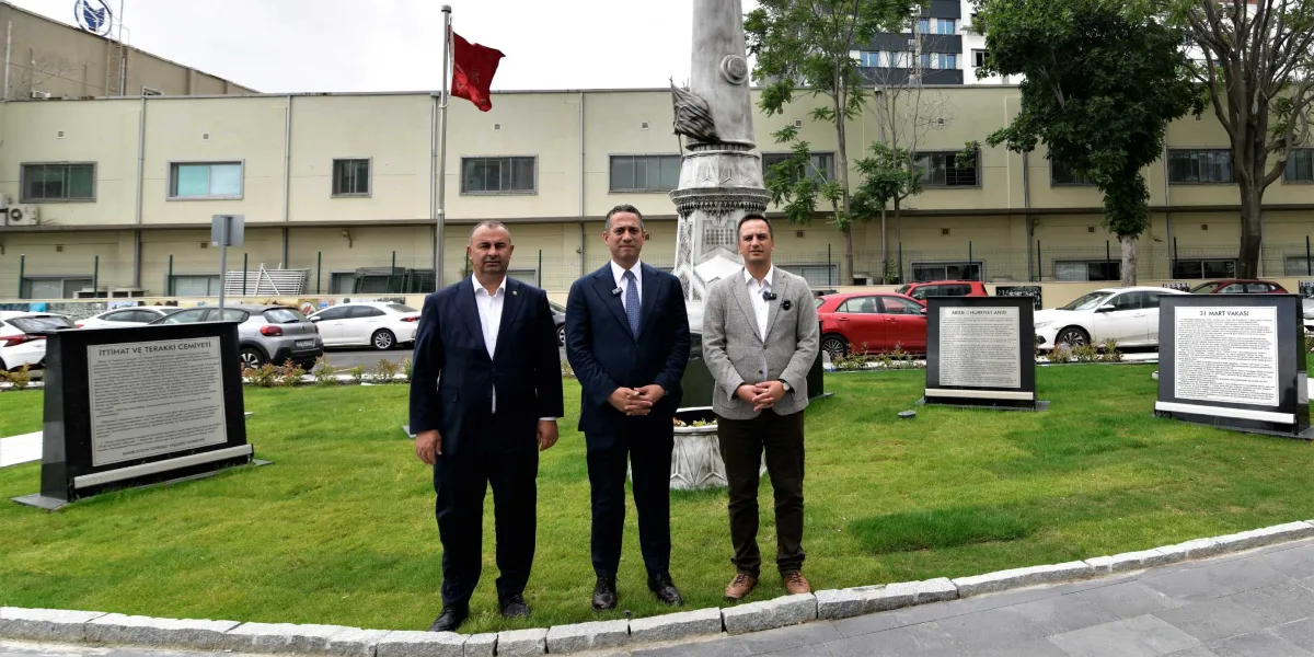 Bornova Abide-i Hürriyet Anıtı önünde yan yana duran üç erkek; arka planda yeşil alan, park halindeki araçlar ve üzerinde bilgilendirme yazıları bulunan iki adet siyah mermer anıt panosu yer alıyor.