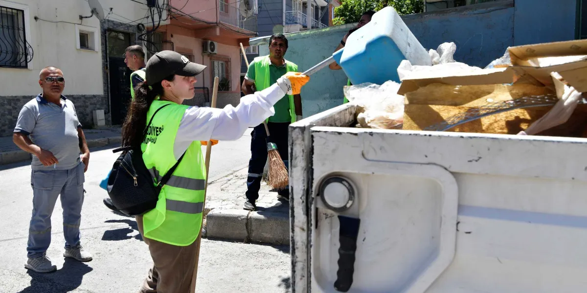 Üzerlerinde neon sarı yelekler bulunan belediye temizlik görevlileri, bir sokak üzerinde çöp toplama çalışması yapıyor. Ön planda, elinde mavi bir çöp küreği tutan kadın görevli, atıkları çöp kamyonunun kasasına boşaltıyor. Arka planda ise elinde süpürge tutan bir erkek görevli ve sokağın kenarında duran başka bir çalışan yer alıyor.