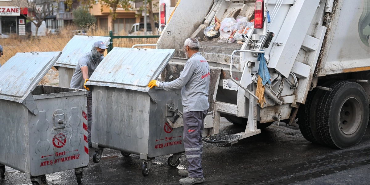 Bornova Belediyesi üniformalı iki temizlik işçisi, sokaktaki metal çöp konteynerlerini çöp kamyonuna boşaltıyor. İşçilerden biri konteynerin kapağını açarken diğeri konteyneri kamyonun arkasındaki kaldırma mekanizmasına yanaştırıyor. Konteynerlerin üzerinde kırmızı yazıyla 