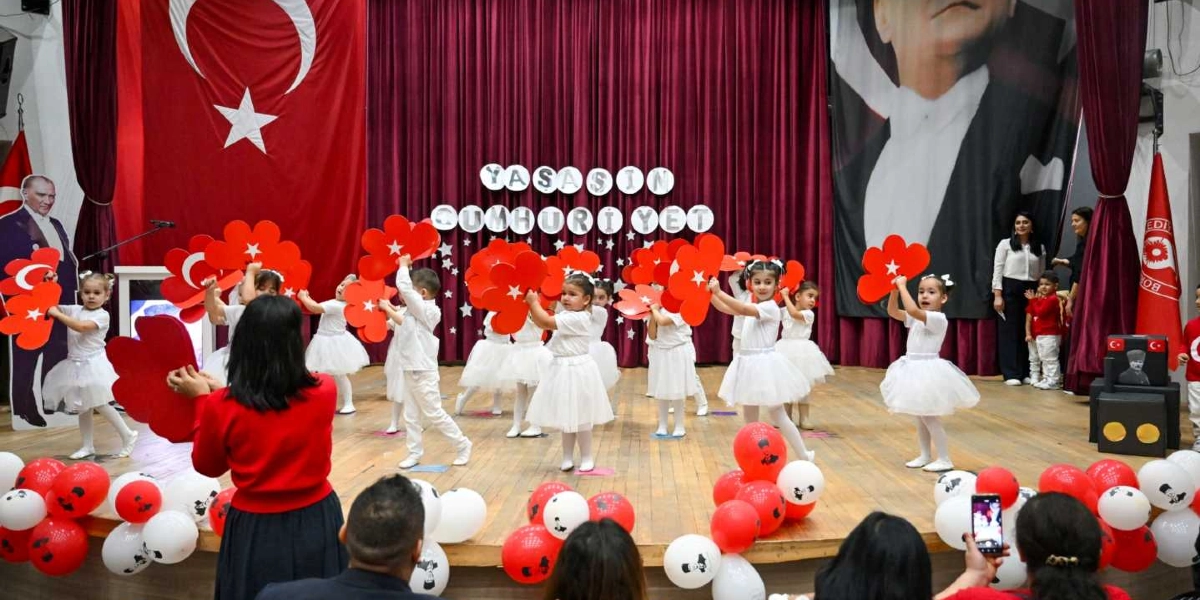 Kırmızı perdeli bir sahnede, beyaz kıyafetler giymiş anaokulu öğrencileri ellerinde kırmızı çiçek şeklinde süslerle gösteri yapıyor. Sahnenin arka planında büyük bir Türk bayrağı, Mustafa Kemal Atatürk'ün portresi ve 