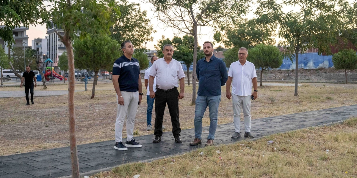 Açık havada, park benzeri bir alanda yan yana duran dört erkek, çevrelerini inceliyor. Ön planda, siyah kauçuk zeminli bir yürüyüş yolunda duran grupta; lacivert polo yaka tişörtlü, beyaz pantolonlu bir kişi, yanında beyaz kısa kollu gömlek ve siyah pantolon giyen bıyıklı bir kişi, onun yanında lacivert gömlek ve kot pantolonlu bir kişi ve en sağda beyaz kısa kollu gömlek ile açık renk pantolon giyen bir kişi yer alıyor. Arka planda ise ağaçlar, çocuk oyun parkı ve uzakta binalar görülüyor.