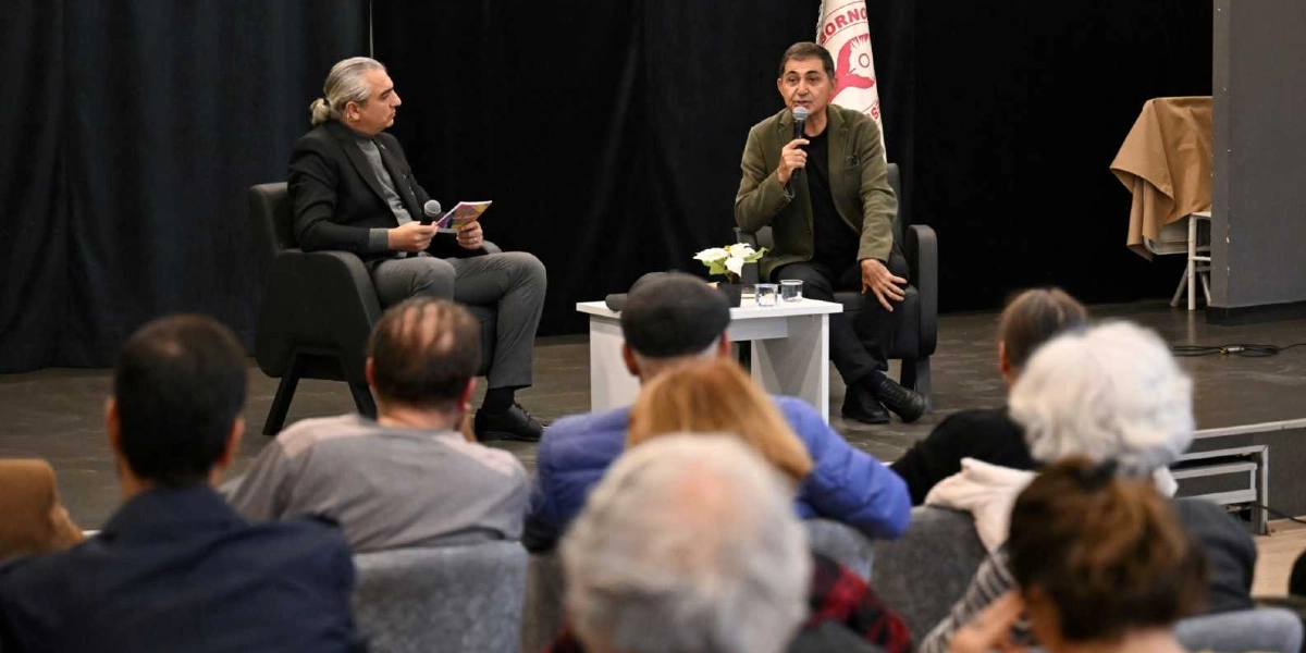 Siyah bir sahne perdesi önünde, Bornova Belediyesi logosunun yer aldığı bir bayrağın yanında, iki kişi koltuklarda oturarak söyleşi gerçekleştiriyor. Sağ tarafta oturan kişi elinde mikrofonla konuşurken, sol tarafta oturan ve saçlarını arkadan toplamış olan kişi elinde bir not defteriyle onu dinliyor. İkili arasında küçük bir sehpa üzerinde çiçek ve su bardakları bulunuyor. Ön planda ise söyleşiyi izleyen dinleyicilerin sırtları görünüyor.