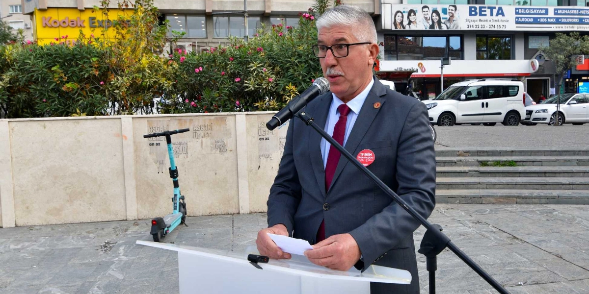 Gri takım elbiseli, bordo kravatlı ve gözlüklü bir beyefendi, açık havada bir kürsüde mikrofon başında konuşma yapıyor. Yakasında 19 Ekim Muhtarlar Günü yazılı kırmızı bir rozet bulunan kişi, elindeki küçük bir kağıttan notlarını okuyor. Arka planda Bornova'daki bir meydanda ağaçlar, park halindeki araçlar ve çevredeki iş yerlerinin tabelaları görülüyor.