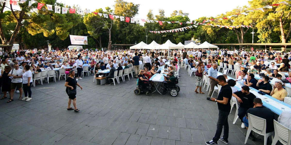 Ağaçlarla çevrili geniş bir açık alanda, uzun masaların etrafında toplanmış kalabalık bir insan grubu. Masaların üzeri mavi beyaz örtülerle kaplıdır ve arka planda beyaz çadırlar kuruludur. Alanın üzerinde Türk bayrakları ve çeşitli flamalarla süslenmiş ipler asılıdır. Arka tarafta 