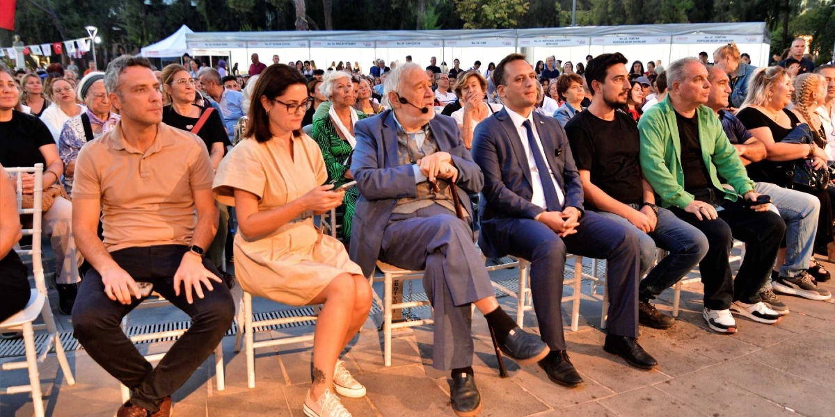 Açık havada düzenlenen bir etkinlikte, ön sırada yan yana oturan İlber Ortaylı ve beraberindeki protokol üyeleri ile izleyiciler görülüyor. İlber Ortaylı, mavi takım elbisesi ve elinde tuttuğu bastonuyla dikkat çekerken, yanında bej rengi bir elbise giyen bir kadın, lacivert takım elbiseli bir erkek ve diğer katılımcılar yer alıyor. Arka planda ise beyaz çadırlar ve kitap stantlarının bulunduğu bir fuar alanı seçiliyor.
