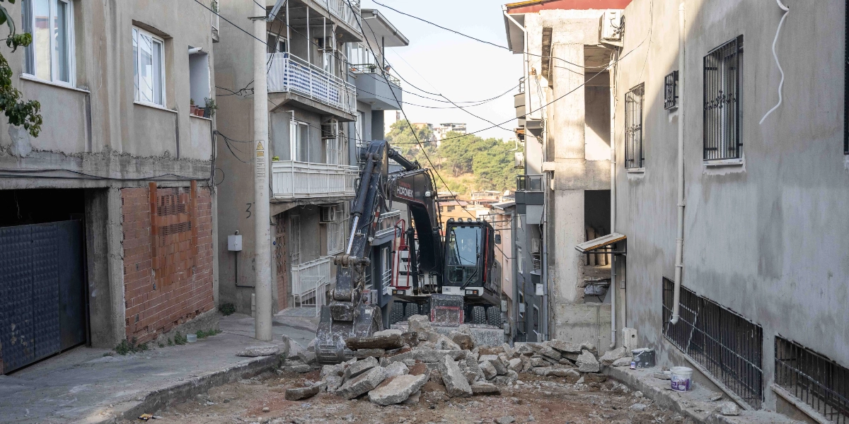Bornova'da dar bir sokak arasında çalışan siyah renkli bir Hidromek iş makinesi, kazı alanındaki beton parçalarını temizliyor. Sokağın iki yanında çok katlı apartmanlar bulunuyor ve zemin seviyesinde yol yapım çalışmaları devam ediyor.