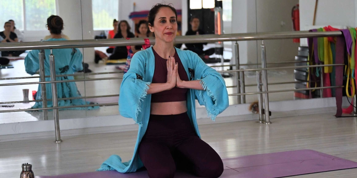 Bordo renkli spor kıyafetleri giymiş bir kadın, üzerinde açık mavi, uzun kollu bir hırka ile yoga matı üzerinde diz çökmüş oturuyor. Elleri göğüs hizasında birleştirilmiş dua pozisyonunda duruyor ve hafifçe gülümseyerek karşıya bakıyor. Arka planda, bir spor salonunda yoga yapan diğer kadınların yansımasının görüldüğü boydan boya bir ayna ve duvarda asılı renkli direnç bantları yer alıyor.
