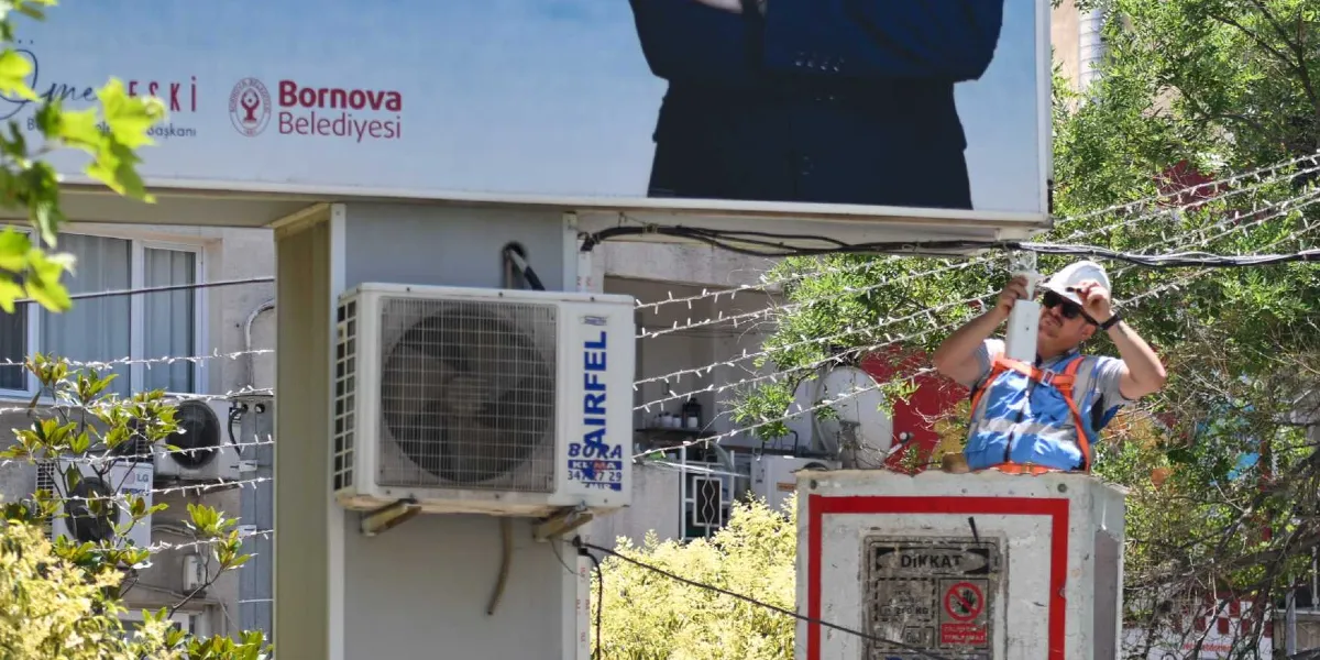 Beyaz baretli, güneş gözlüklü ve reflektörlü yelek giyen bir teknisyen, sepetli bir vinç üzerinde yükselerek büyük bir reklam panosunun altına teknik bir cihaz montajı yapıyor. Reklam panosunun üzerinde Bornova Belediyesi logosu ve bir kişinin silüeti yer alırken, panonun hemen altında bir Airfel marka klima dış ünitesi bulunuyor.