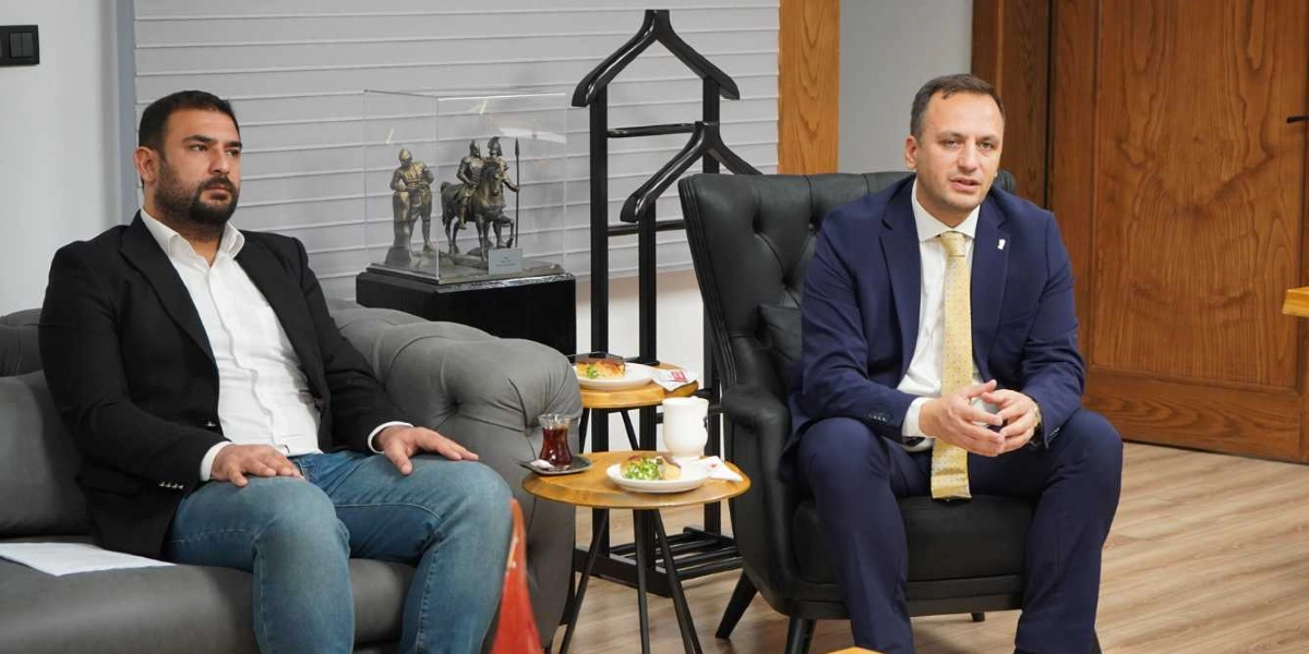 Modern bir ofis ortamında, gri bir koltukta oturan sakallı ve siyah ceketli bir erkek ile yanında siyah deri bir koltukta oturan, lacivert takım elbiseli ve sarı kravatlı bir erkek sohbet ediyor. İkilinin arasında küçük ahşap sehpalar üzerinde çay ve atıştırmalıklar bulunuyor; arka planda ise şeffaf bir kutu içerisinde atlı asker figürleri ve bir askılık yer alıyor.
