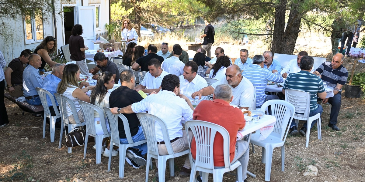 Ağaçların gölgesinde, dış mekanda kurulmuş uzun masaların etrafında bir araya gelmiş kalabalık bir grup insan yemek yiyor. Beyaz plastik sandalyelerde oturan katılımcılar, Bornova Belediyesi logolu örtülerin serili olduğu masalarda sohbet ederek vakit geçiriyor. Arka planda beyaz bir yapı ve ağaçlık bir arazi görülüyor.