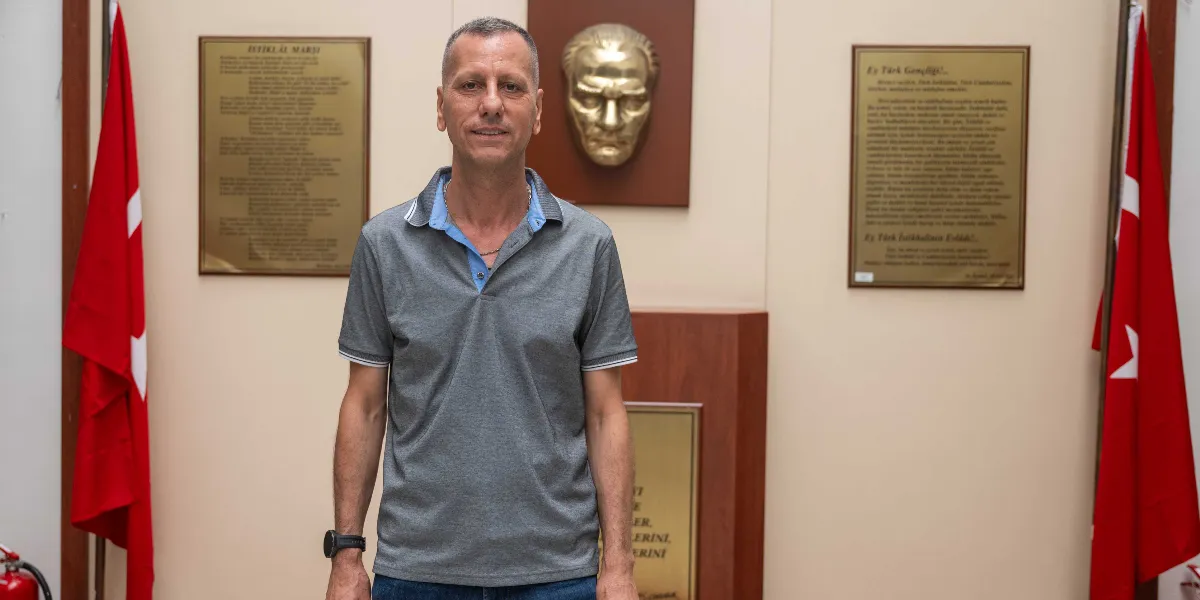 Gri polo yaka tişört giymiş, kısa saçlı bir erkek, arka planda Atatürk rölyefi, İstiklal Marşı ve Gençliğe Hitabe metinlerinin bulunduğu, iki yanında Türk bayraklarının yer aldığı bir odada ayakta duruyor.