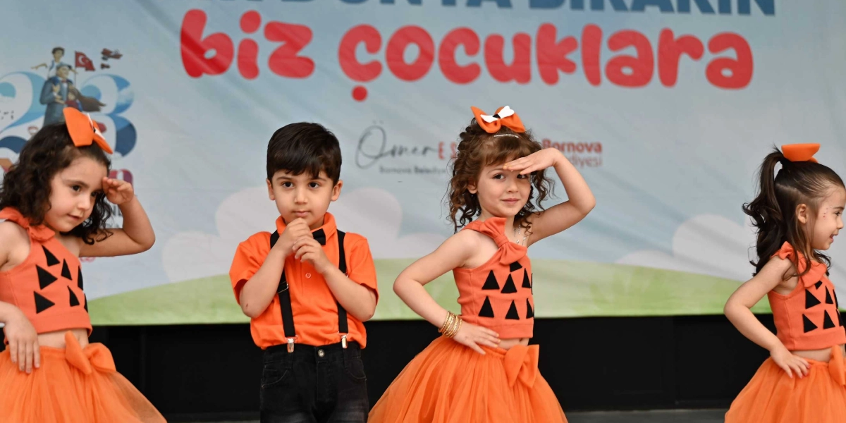 23 Nisan gösterisi için turuncu kostümler giymiş dört çocuk sahnede dans ediyor. Kız çocuklar üzerinde siyah üçgen desenli turuncu büstiyer ve tül etekler, saçlarında ise turuncu fiyonklar bulunuyor. Erkek çocuk ise turuncu bir tişört, siyah pantolon ve askı takıyor. Çocukların arkasındaki panoda 