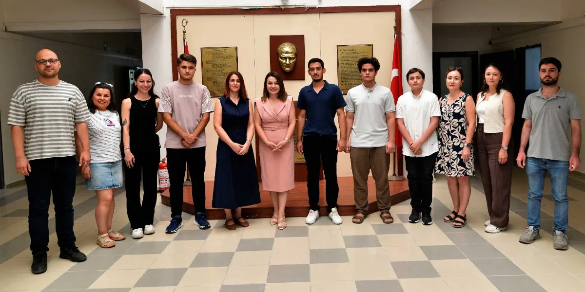 Arka planda Atatürk büstü ve iki yanında Türk bayraklarının bulunduğu bir salonda, yan yana dizilmiş 13 kişilik bir grup insan poz veriyor. Grupta farklı yaş ve tarzlarda giyinmiş kadın ve erkekler yer alıyor; bazıları günlük kıyafetler, bazıları ise daha resmi elbiseler tercih etmiş. Hepsi gülümseyerek doğrudan kameraya bakıyor.