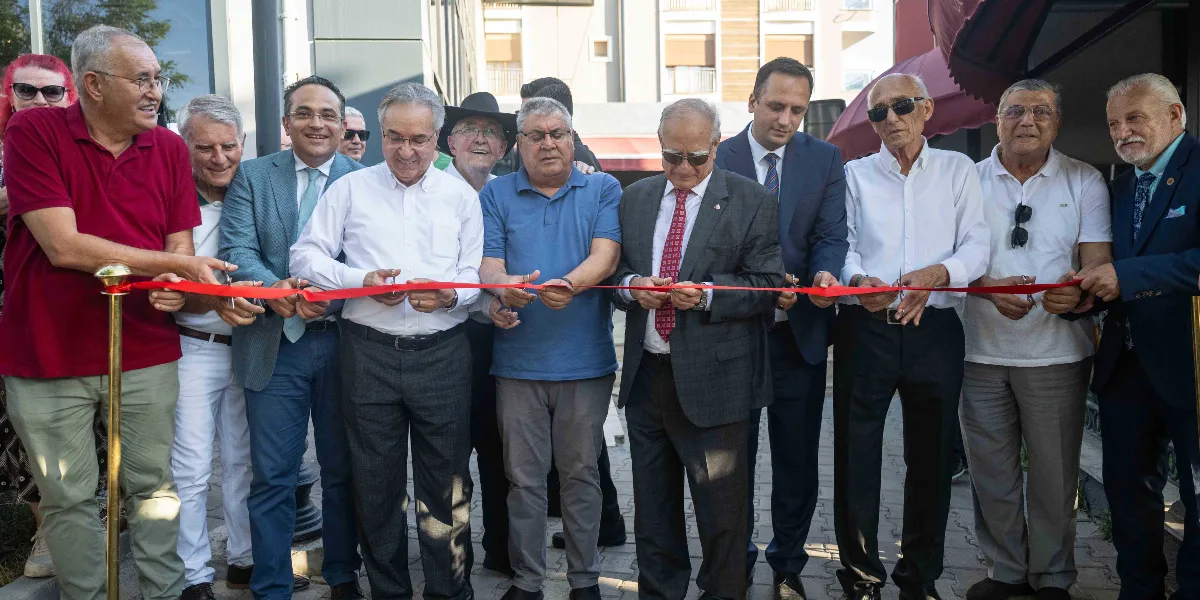 Bornova'da bir tesisin açılış töreninde, aralarında Bornova Belediye Başkanı Mustafa İduğ'un da bulunduğu bir grup yetkili ve davetli, ellerinde tuttukları kırmızı kurdeleyi kesmek üzere yan yana dizilmiş duruyor. Açık havada gerçekleşen törende, takım elbiseli ve günlük kıyafetli erkeklerden oluşan grup, kurdeleyi kesmek için hazırlık yaparken gülümsüyor.