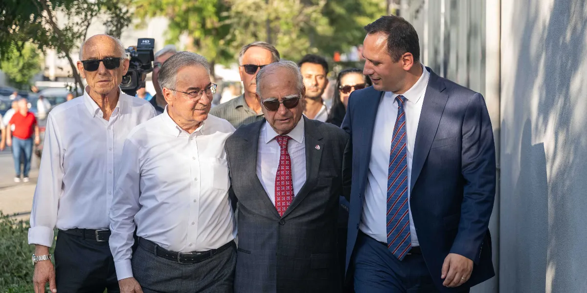 Açık havada yan yana yürüyen dört erkekten oluşan bir grup. En solda güneş gözlüklü ve beyaz gömlekli bir kişi, yanında gözlüklü ve yine beyaz gömlekli bir bey, ortada koyu renk takım elbiseli ve kırmızı desenli kravatlı bir bey, en sağda ise lacivert takım elbiseli ve çizgili kravatlı bir bey yer alıyor. Arka planda ise bulanık bir şekilde diğer katılımcılar ve ağaçlar görünüyor.