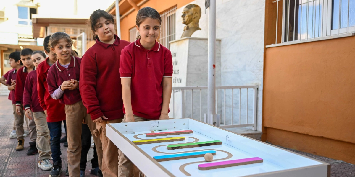 Okul bahçesinde bordo polo yaka tişörtlü ve bej pantolonlu bir grup öğrenci, önlerinde duran ahşap bir labirent oyununu sırayla oynamak için kuyruk oluşturmuş bekliyor. En öndeki öğrenci, üzerinde renkli engellerin bulunduğu labirent masasını elleriyle tutarken, arkasındaki diğer öğrenciler merakla oyunu izliyor. Arka planda ise üzerinde Atatürk büstü bulunan bir okul binası yer alıyor.