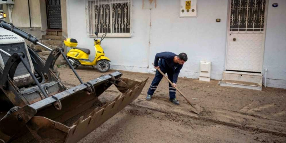 Bornova Belediyesi'ne ait bir iş makinesi, sel sonrası çamurla kaplanan sokakta temizlik çalışması yapıyor. İş kıyafetleri giymiş bir belediye çalışanı, elindeki kürekle çamurları temizlerken, arka planda beyaz bir binanın önünde park edilmiş sarı bir motosiklet görülüyor.