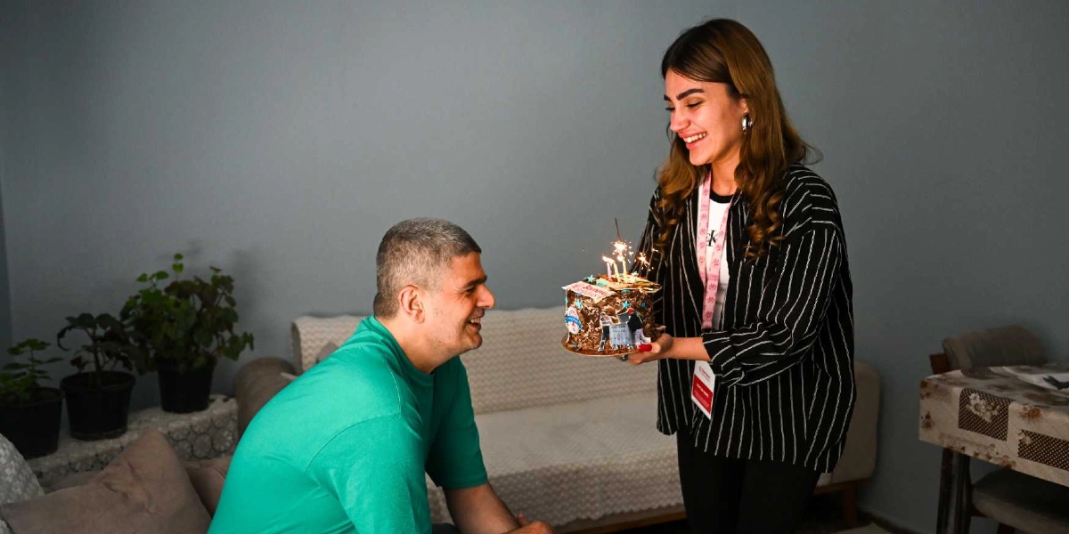 Yeşil tişörtlü, kısa saçlı bir adam, karşısında duran ve üzerinde yanan mumlar bulunan küçük bir pastayı kendisine uzatan, siyah beyaz çizgili gömlekli genç bir kadına gülümseyerek bakıyor. Kadının boynunda bir kimlik kartı asılı ve arka planda gri bir duvarın önünde saksı bitkileri ile bir koltuk bulunuyor.