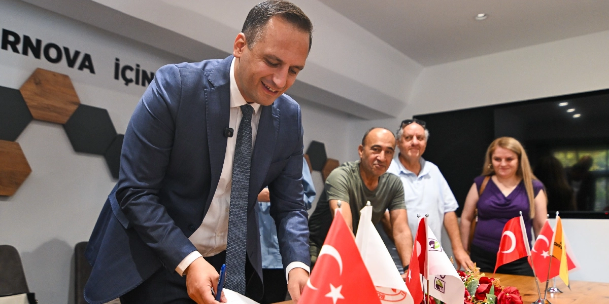 Lacivert takım elbiseli bir erkek, üzerinde Bornova Belediyesi logosu bulunan bir belgenin altına imza atarken gülümsüyor. Masanın üzerinde küçük Türk bayrakları ve kurum flamaları yer alıyor. Arka planda ise üç kişi bu anı izliyor; duvarda ise 