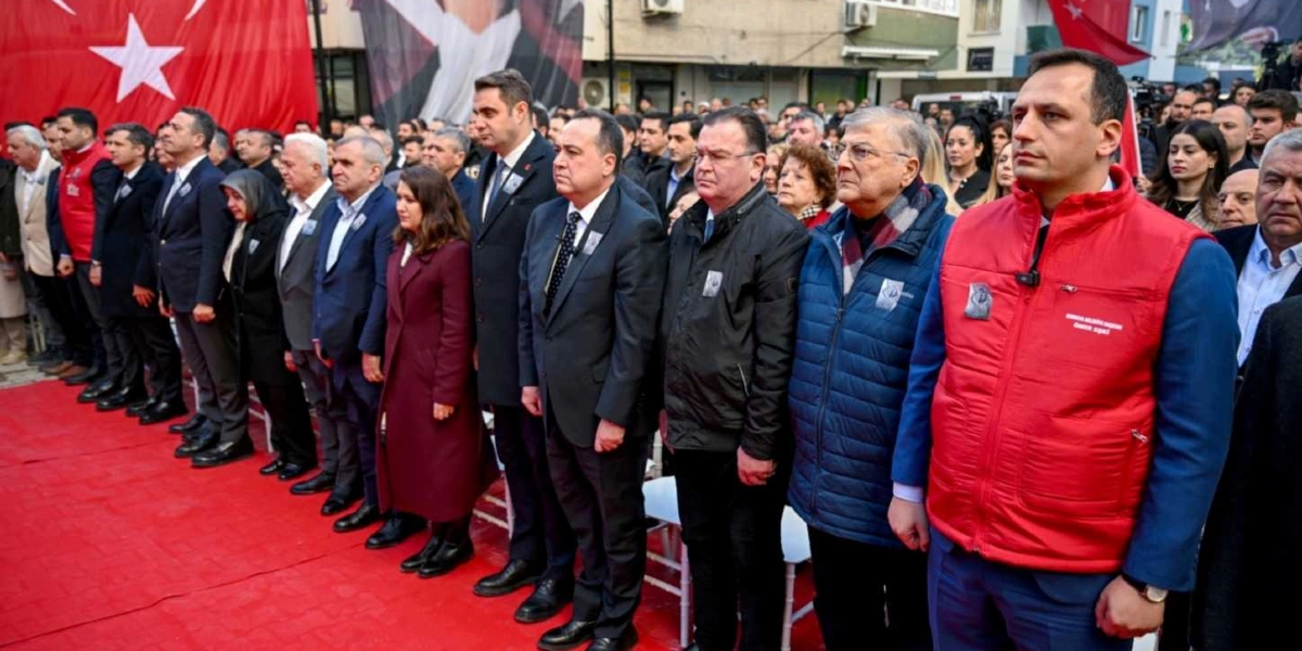 Açık havada, kırmızı bir halı üzerinde yan yana dizilmiş, resmi kıyafetli bir grup insan saygı duruşunda bulunuyor. Grubun en önünde, üzerinde 