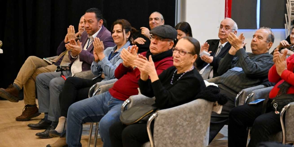 Bir konferans salonunda yan yana oturmuş, alkışlayan bir grup yetişkin izleyici. Ön sırada, siyah kazaklı ve gözlüklü bir kadın, yanında kırmızı kazaklı ve kasketli bir adam, onun yanında ise kot ceketli bir kadın ve mor ceketli bir adam yer alıyor. Arka sıralarda ise takım elbiseli ve günlük kıyafetli diğer katılımcılar dikkatle izleyip alkışlıyor. İzleyicilerin çoğu sahneye doğru dönük ve neşeli bir ifadeyle alkış tutuyor.