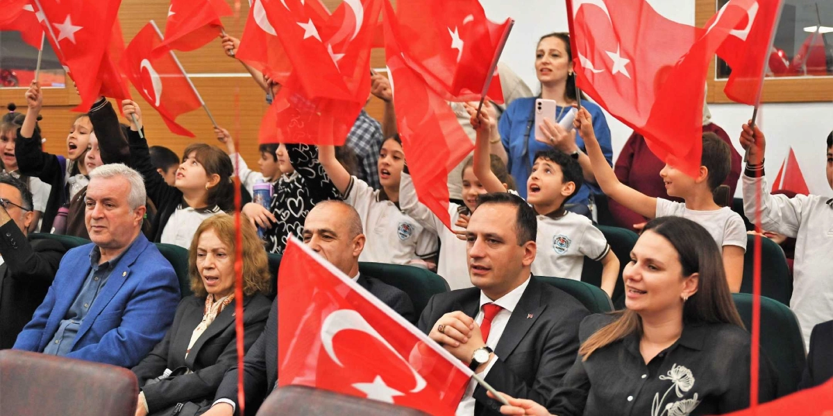 Kapalı bir salonda düzenlenen etkinlikte, ellerinde Türk bayrakları tutan çocuklar ve yetişkinler coşkuyla bir arada oturuyor. Ön sırada takım elbiseli erkekler ve şık giyimli kadınlar yer alırken, arka sıralarda beyaz okul forması giymiş çocuklar heyecanla ellerindeki bayrakları sallıyor. Arka planda bir kadın cep telefonuyla çekim yaparken, genel atmosfer 23 Nisan Ulusal Egemenlik ve Çocuk Bayramı kutlamasının neşesini yansıtıyor.